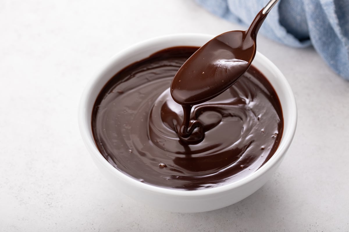 Spoon drizzling chocolate ganache into a white bowl filled with the ganache.