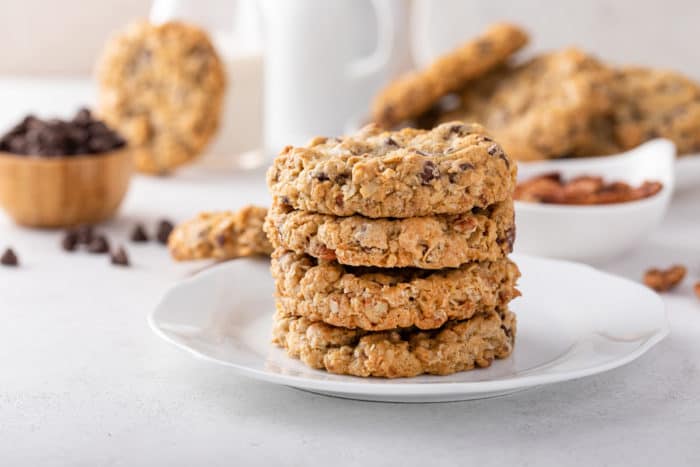 White plate holding a stack of 4 cowboy cookies. Additional cookies and a bowl of chocolate chips are visible in the background.