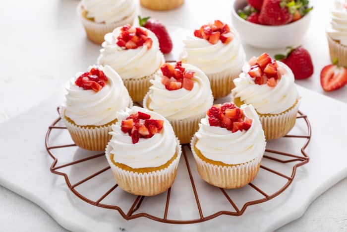 Round wire rack holding seven strawberry shortcake cupcakes topped with fresh strawberries.