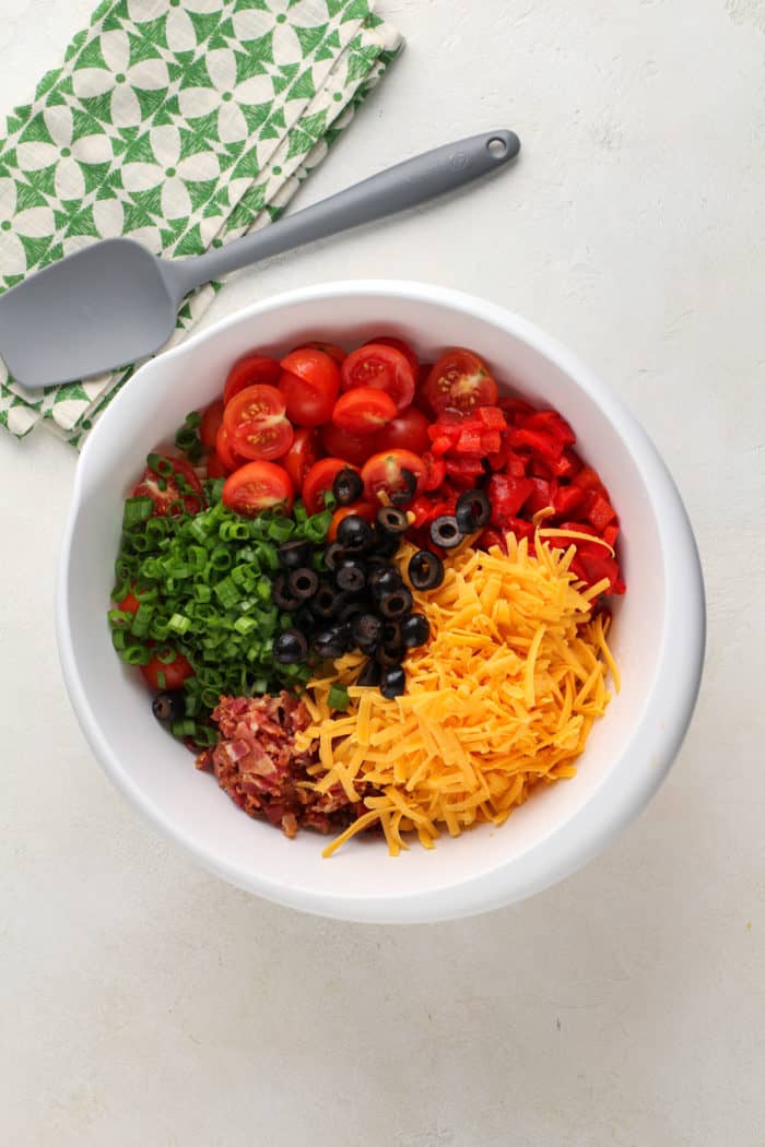 Tomatoes, cheese, bacon, and veggies being added to a white mixing bowl for pasta salad.