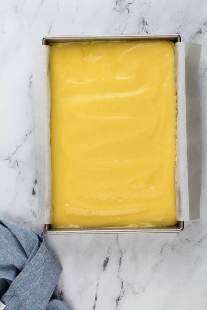 Lemon curd spread onto layered lemon dessert in a parchment-lined baking pan on a marble countertop.