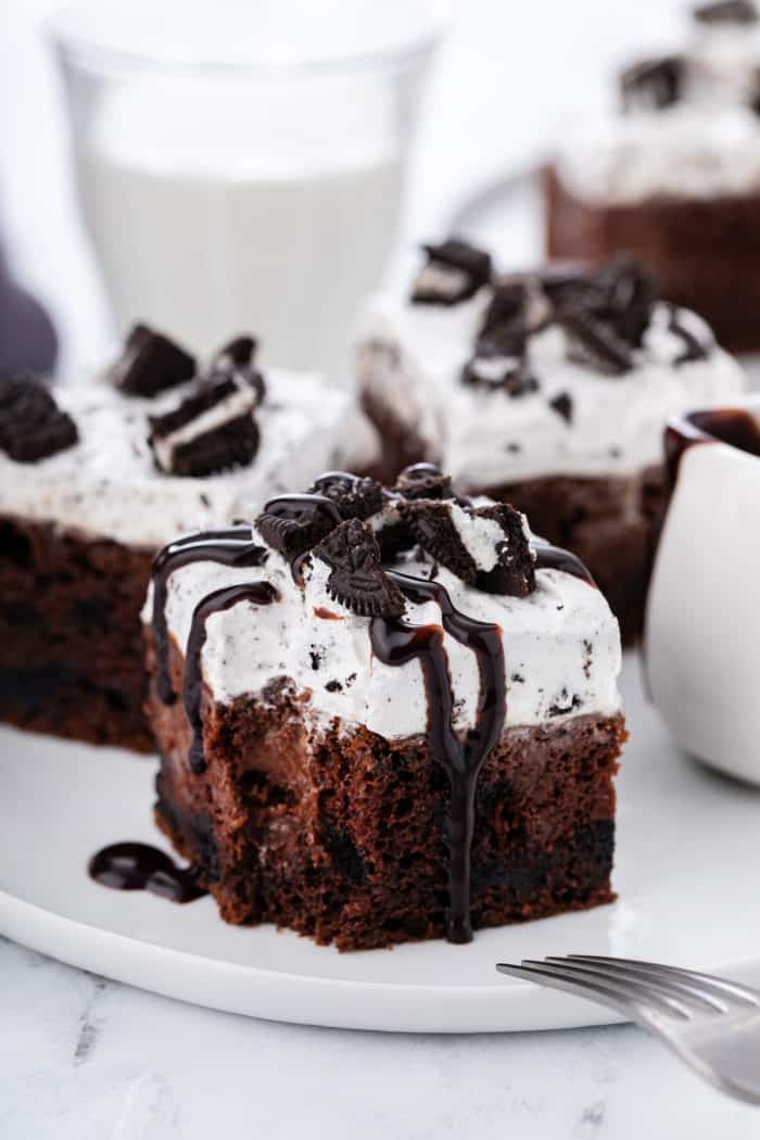 Slice of oreo poke cake drizzled with chocolate sauce, with a bite taken from the corner of the cake.