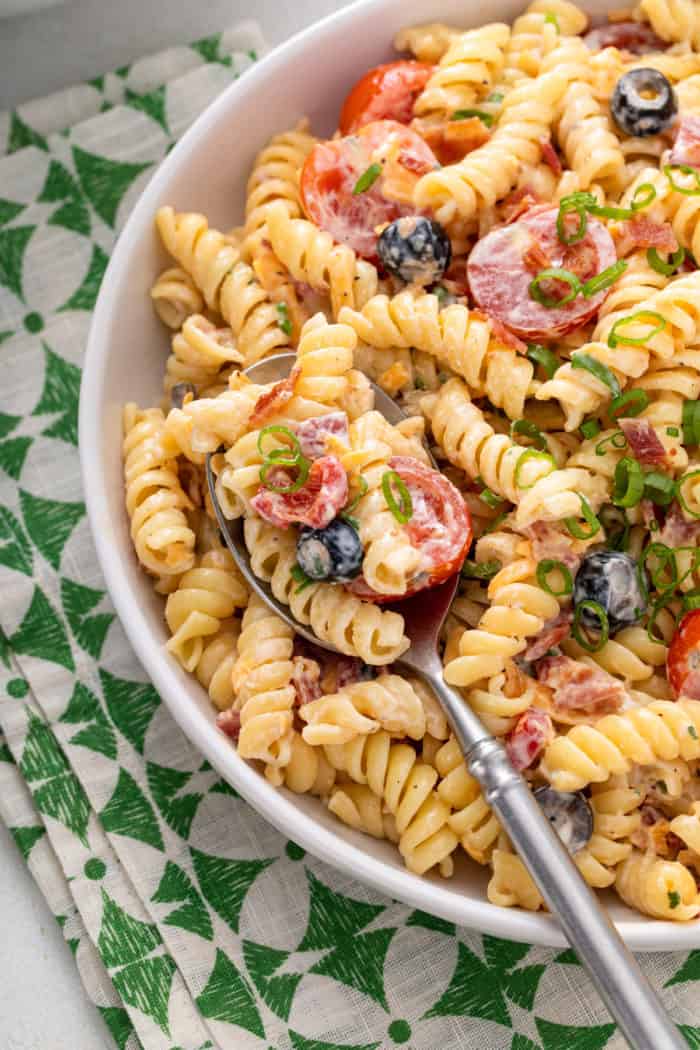 Close up of bacon ranch pasta salad in a white bowl. A silver serving spoon is also in the bowl.