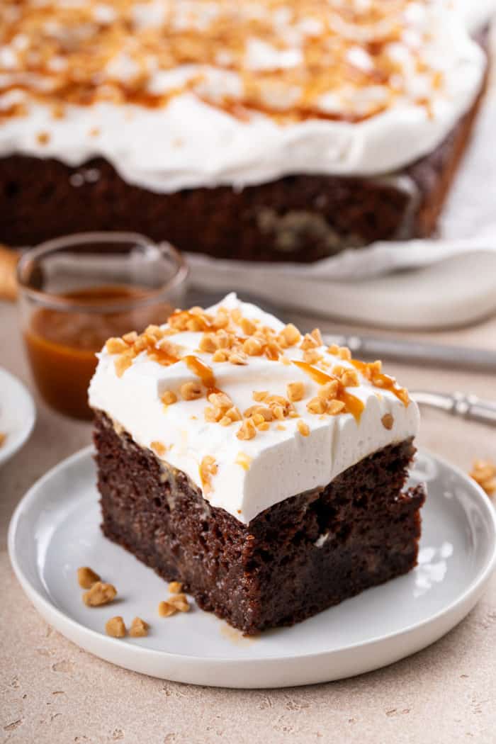 Slice of chocolate toffee poke cake topped with whipped topping and toffee bits on a white plate. The rest of the cake is visible in the background.