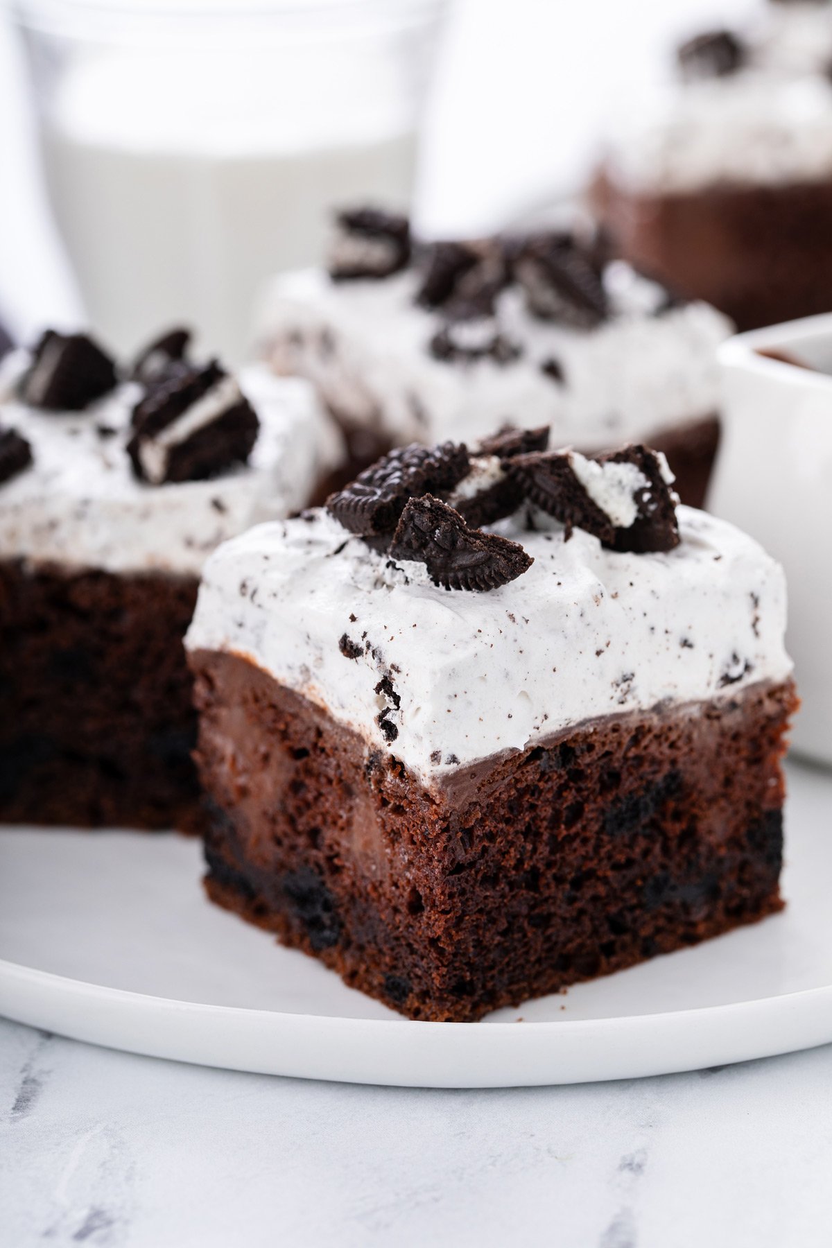 Close up image of a slice of oreo poke cake on a white plate. Two more slices and a glass of milk are visible in the background.