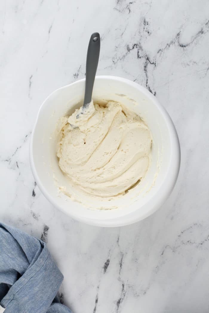 Cream cheese layer for layered lemon dessert being folded with a spatula in a white mixing bowl.