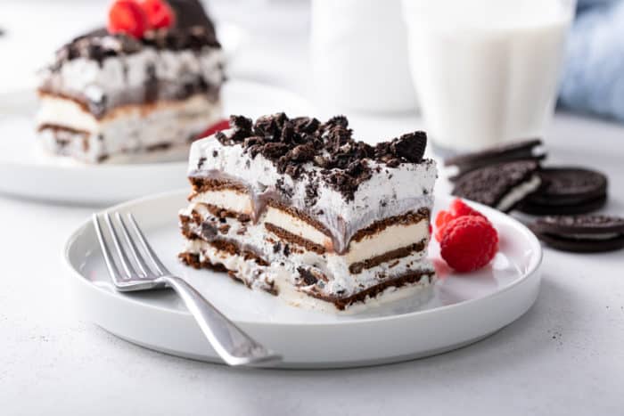 Side view of a slice of ice cream sandwich cake on a white plate. A glass of milk and a second plate are visible in the background.