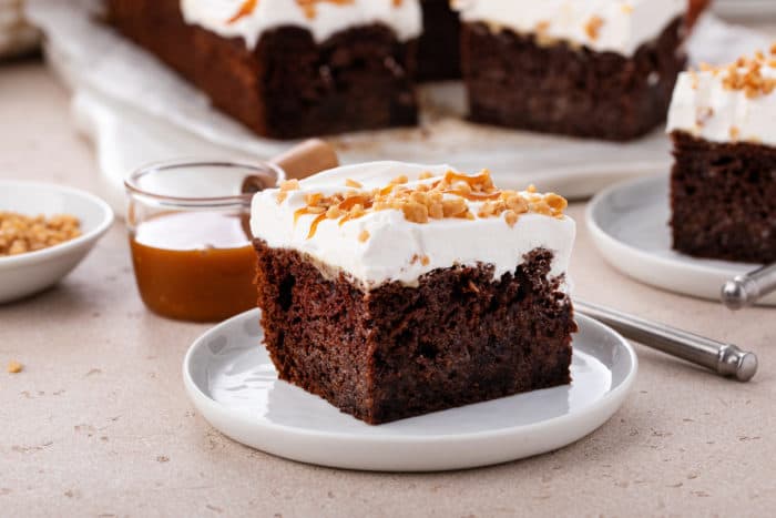 Side view of a slice of chocolate toffee poke cake on a white plate. A bowl of caramel sauce and a second plate of cake are in the background.