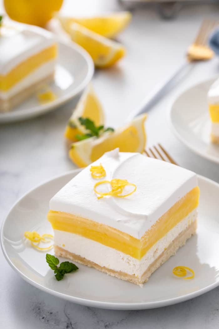 White plate holding a slice of layered lemon dessert that is garnished with lemon zest.