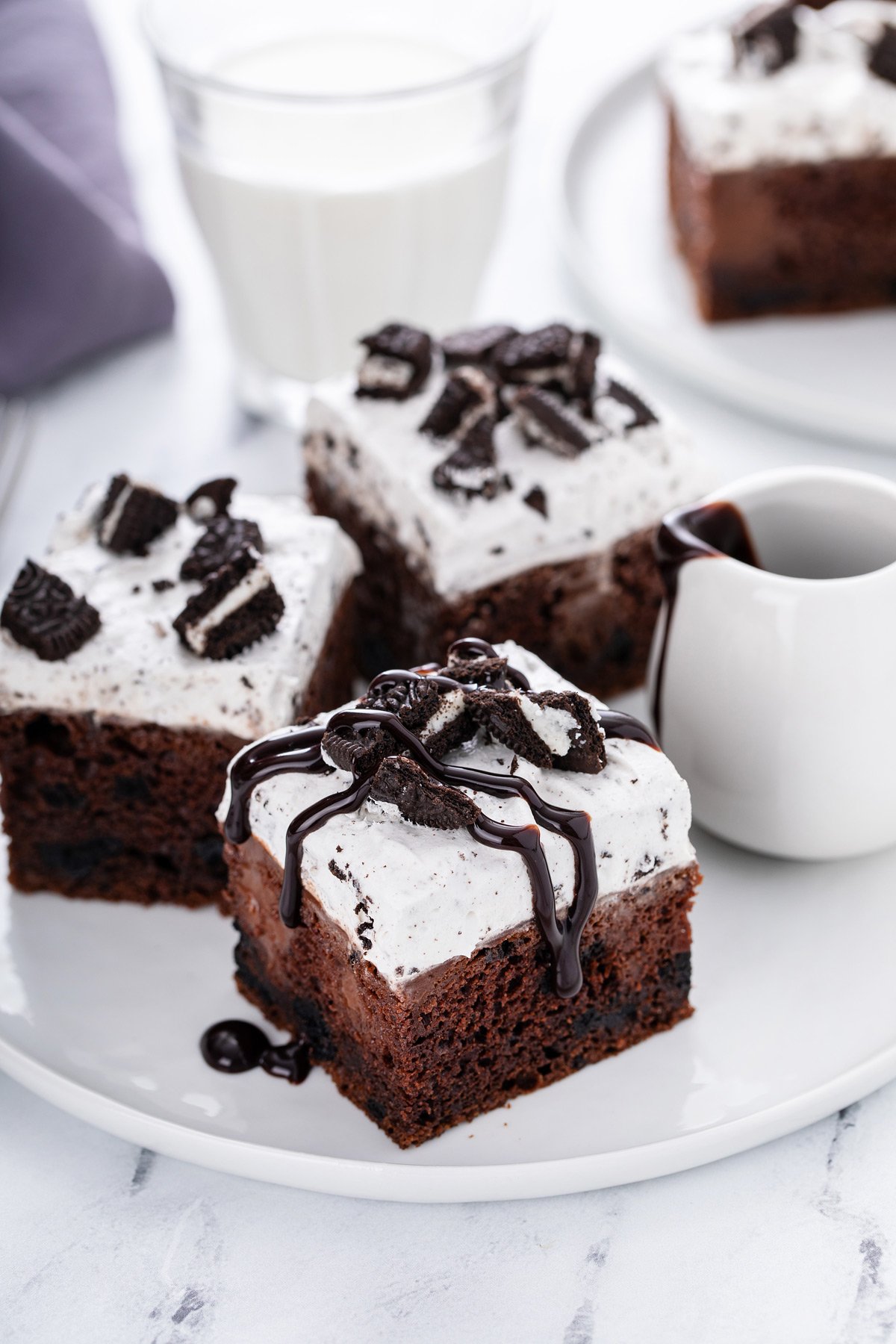 Three slices of oreo poke cake on a white plate. One of the slices is drizzled with hot fudge sauce.