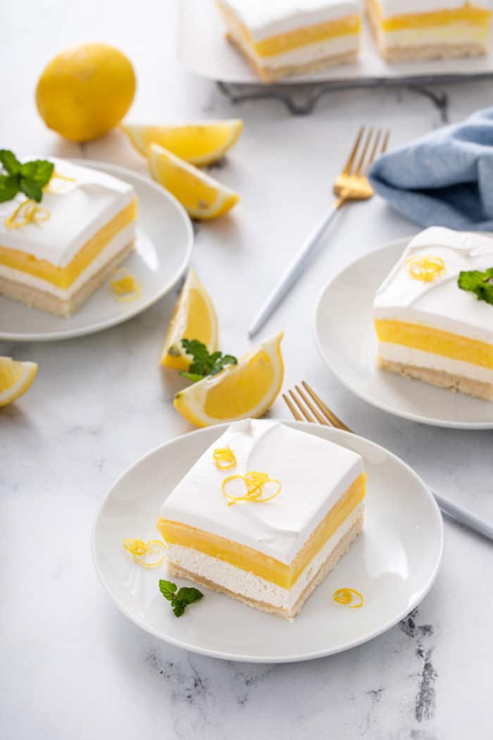 Three white plates, each holding a slice of layered lemon dessert with more dessert in the background.