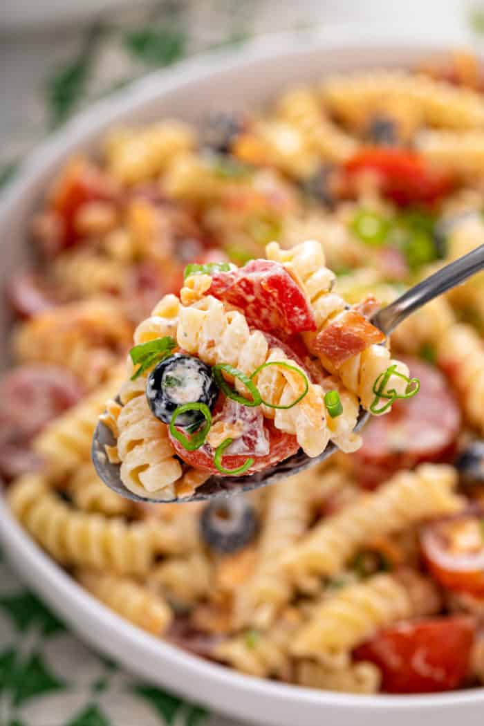 Spoon holding up a big bite of bacon ranch pasta salad, with a bowl of the salad in the background.