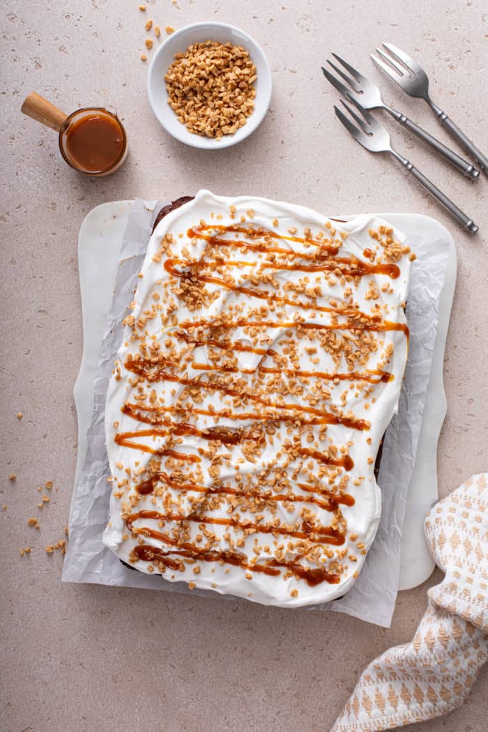 Overhead view of assembled chocolate toffee poke cake, topped with toffee bits and caramel sauce, on a piece of parchment paper.