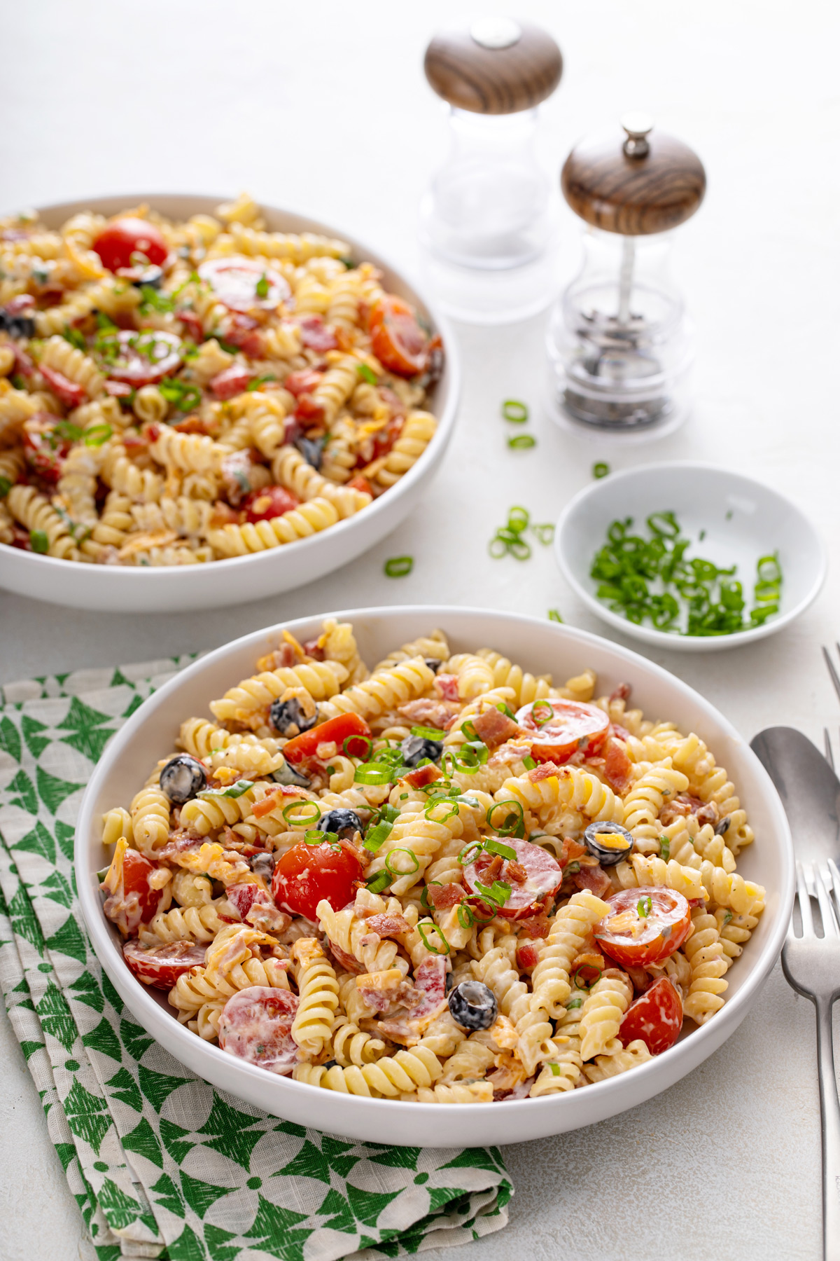 Two white bowls filled with bacon ranch pasta salad, set on a white countertop with a green and white napkin.