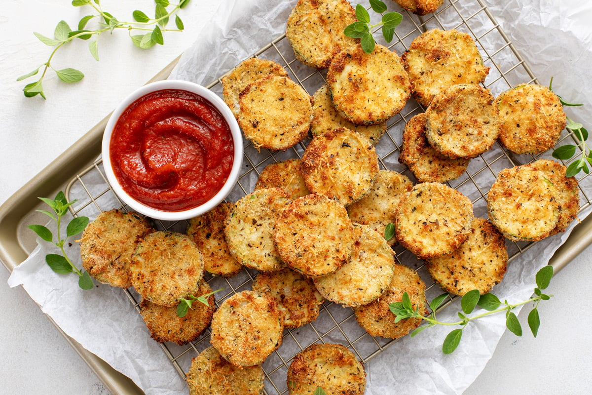 Air fryer zucchini chips and a bowl of marinara sauce set on a parchment-lined baking sheet.