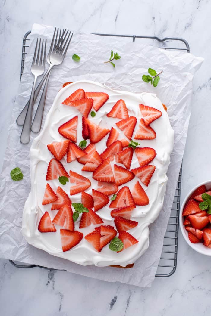 Overhead view of strawberry poke cake topped with cool whip and sliced strawberries on a cooling rack.
