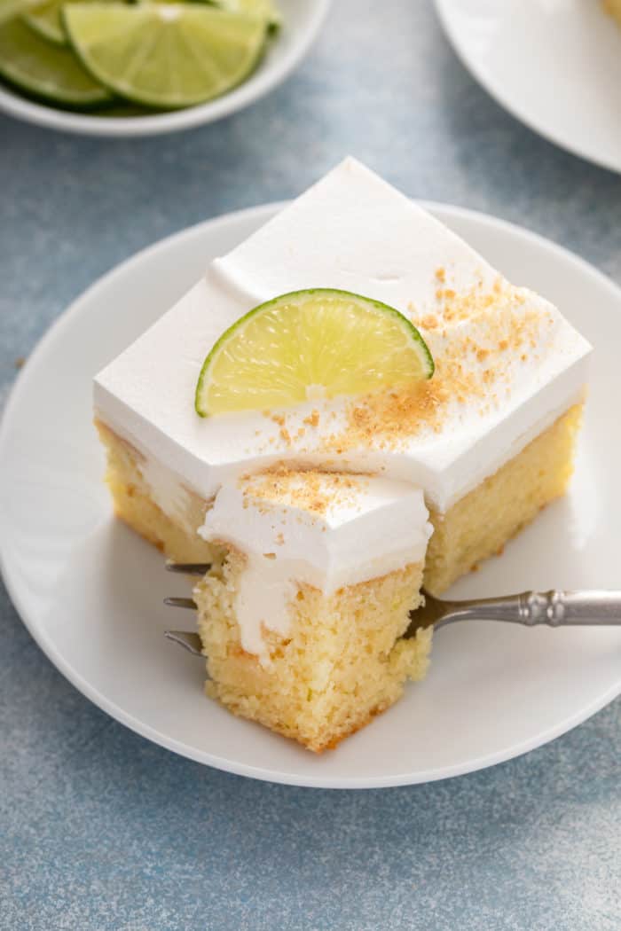 Fork cutting a bite off of the corner of a slice of key lime poke cake on a white plate.