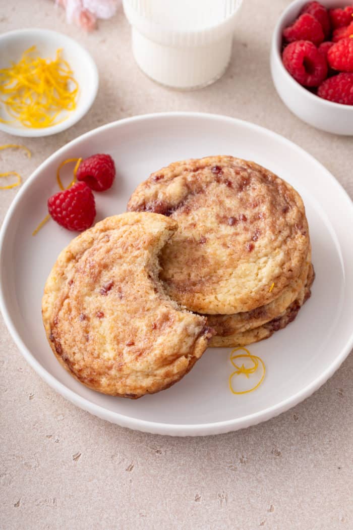 Raspberry lemon cookie with a bite taken out of it leaning against several other cookies on a white plate.