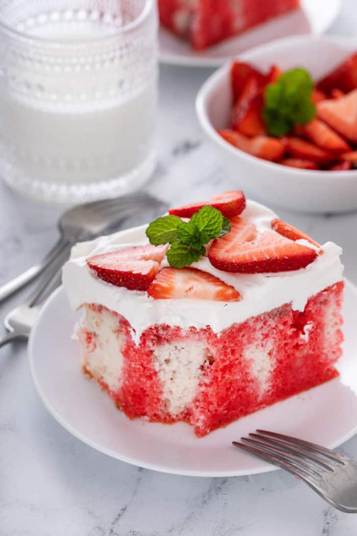 Slice of strawberry poke cake on a white plate. A bite has been taken from the corner of the cake.