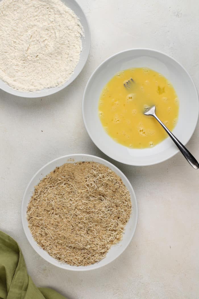 Three shallow bowls, one filled with flour, one filled with beaten eggs, one filled with seasoned breadcrumbs.