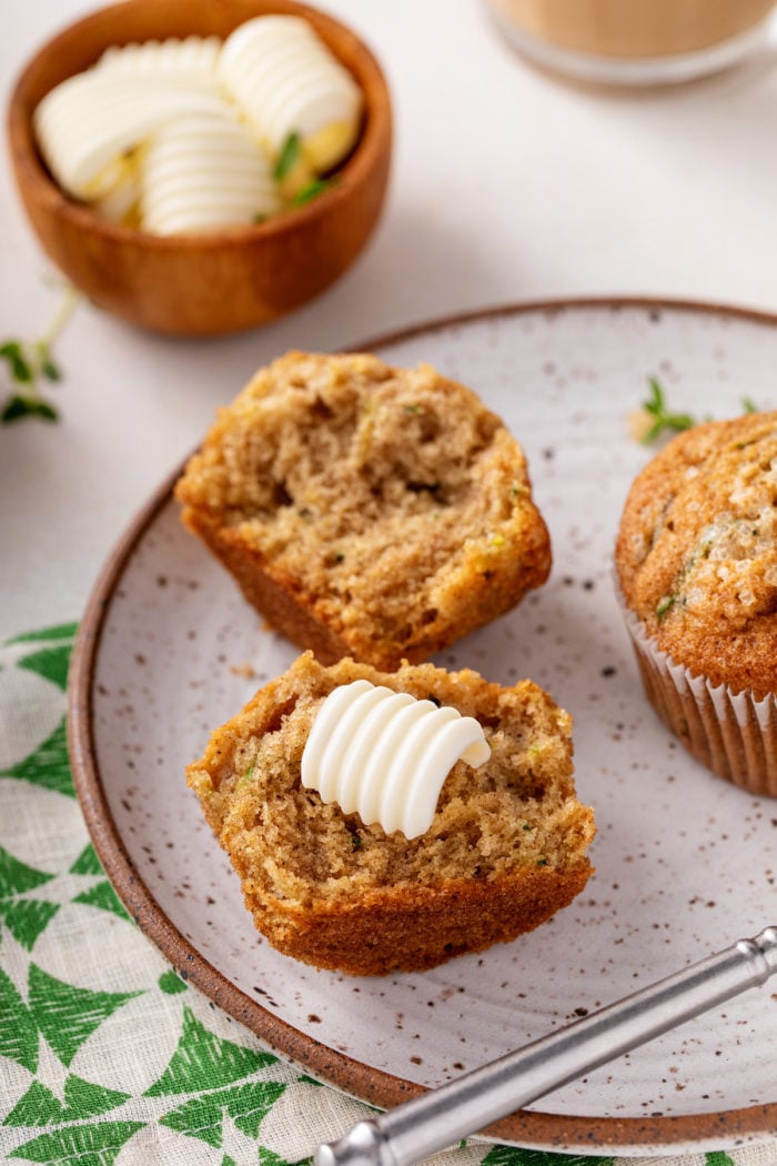 Pat of butter on a zucchini muffin that has been cut in half on a stoneware plate.