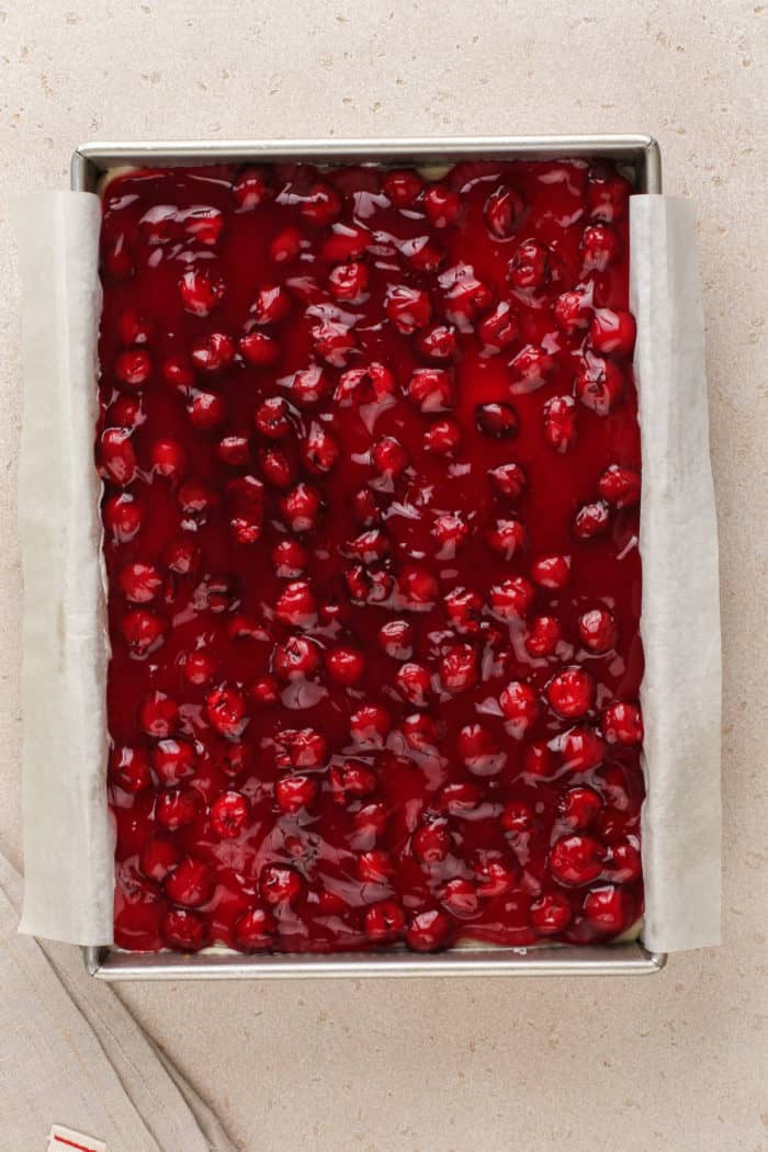 Cherry pie filling spread onto layered cherry cheesecake dessert in a baking pan.