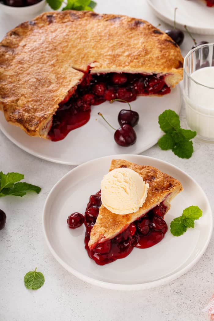 Slice of cherry pie a la mode on a white plate next to a platter holding the rest of the cherry pie.