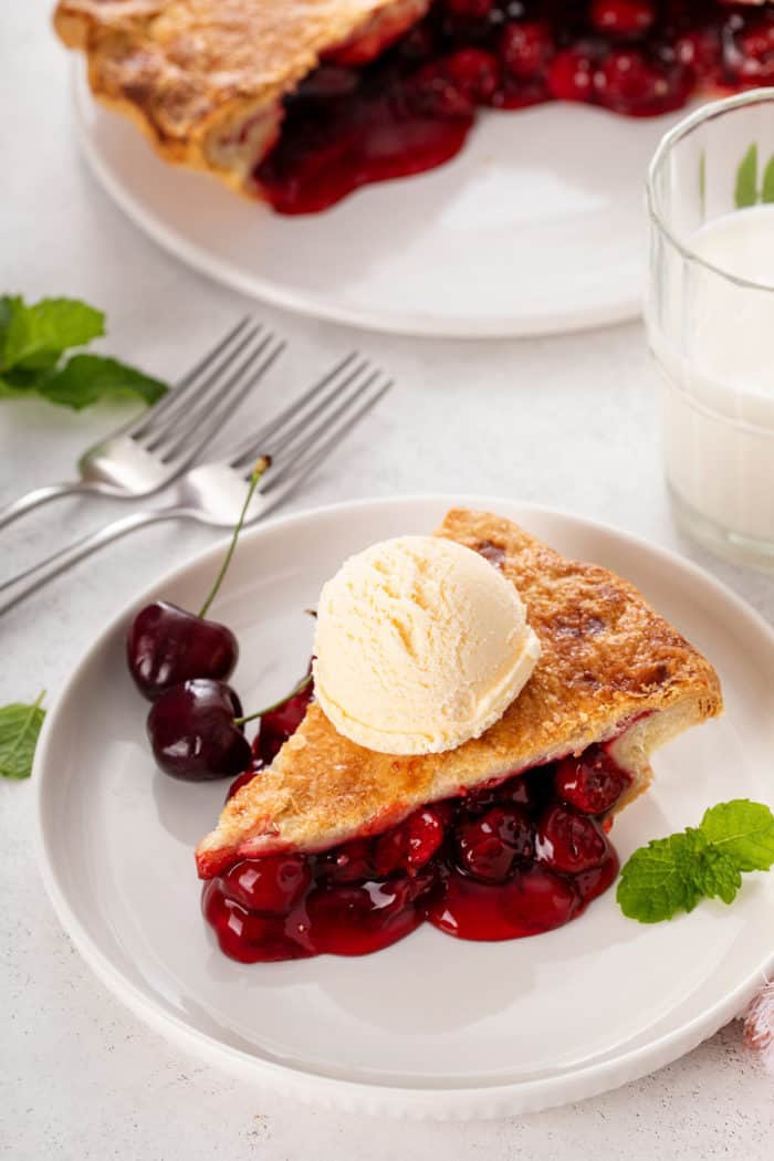 White plate with a slice of cherry pie topped with a scoop of vanilla ice cream.
