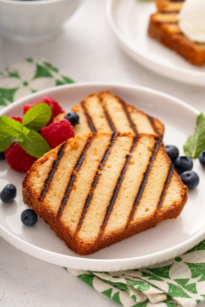 Two slices of grilled pound cake on a white plate with fresh raspberries and blueberries.