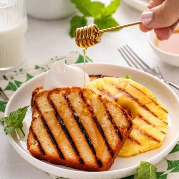 Honey being drizzled over grilled pound cake and grilled pineapple on a white plate.