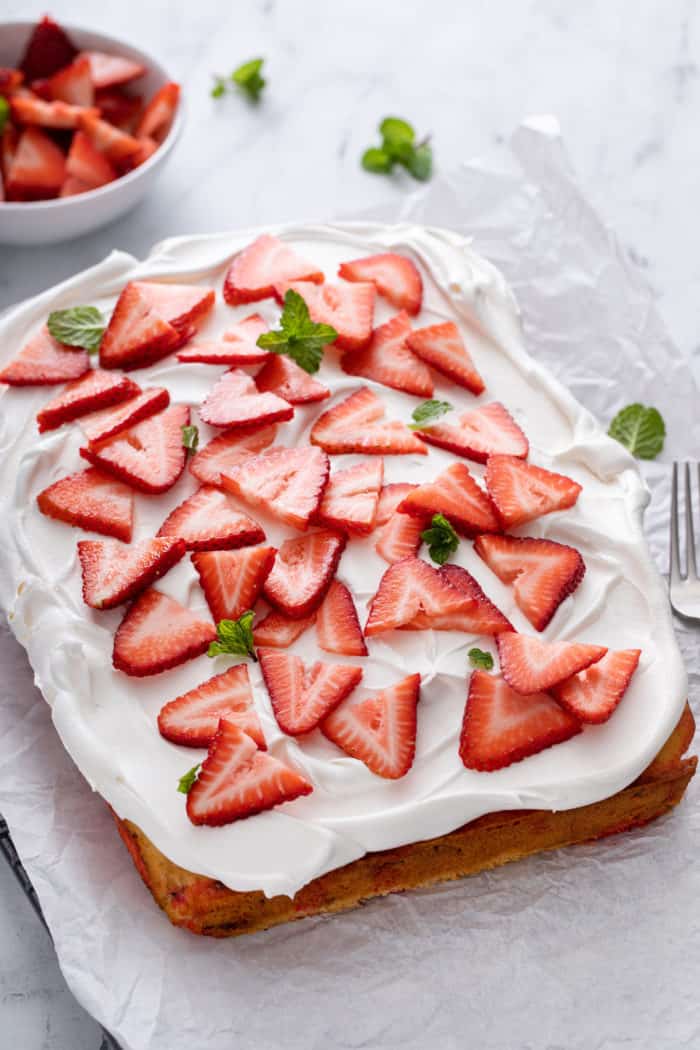 Strawberry poke cake on a piece of parchment paper, topped with whipped topping and sliced fresh strawberries.