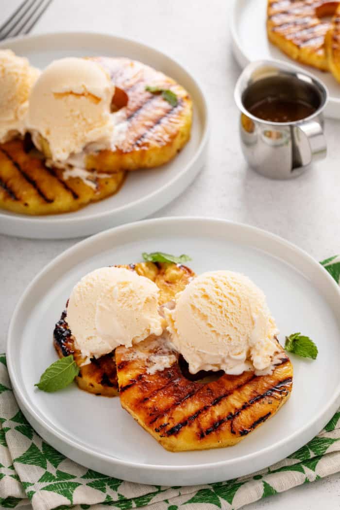 Two white plates, each holding slices of grilled pineapple topped with vanilla ice cream.