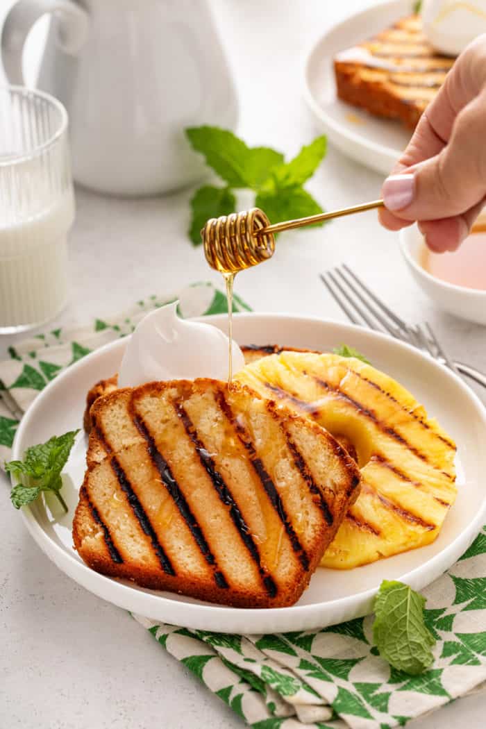 Honey being drizzled over grilled pound cake and grilled pineapple on a white plate.