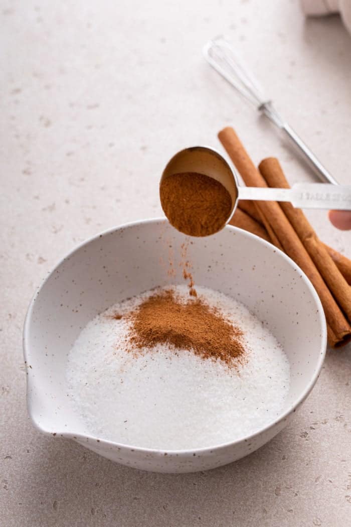 Cinnamon being added to granulated sugar in a white bowl.