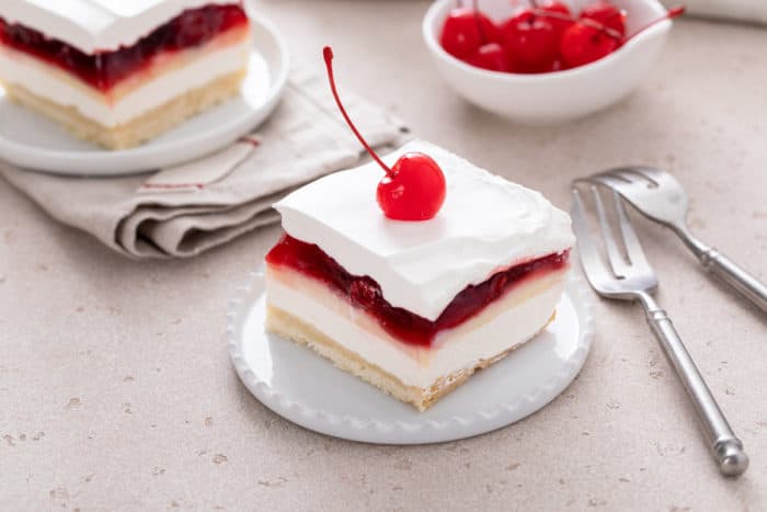 Slice of layered cherry cheesecake dessert topped with a maraschino cherry on a small white plate.
