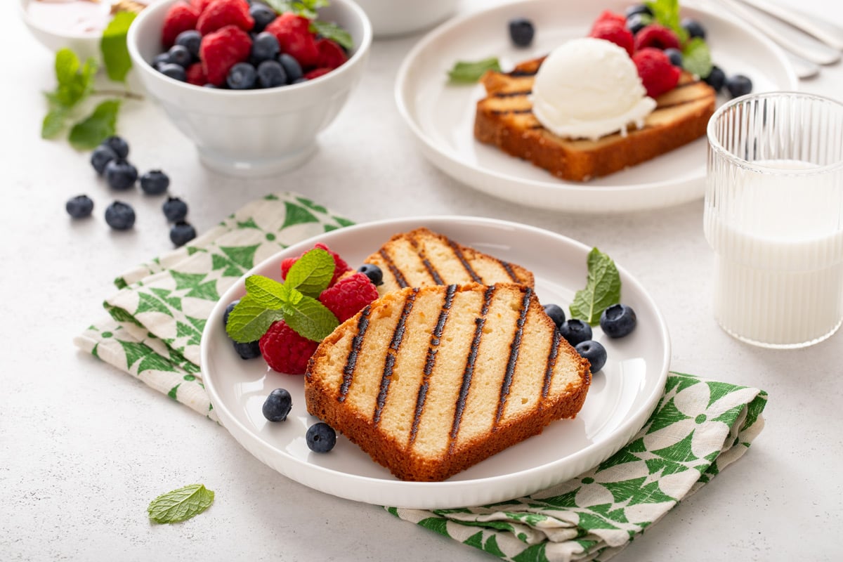 Two slices of grilled pound cake on a white plate. A second plate is visible in the background, holding a slice of grilled pound cake topped with vanilla ice cream.