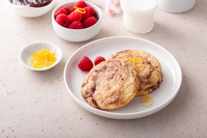 White plate holding several raspberry lemon cookies. A bowl of raspberries and a glass of milk are in the background.