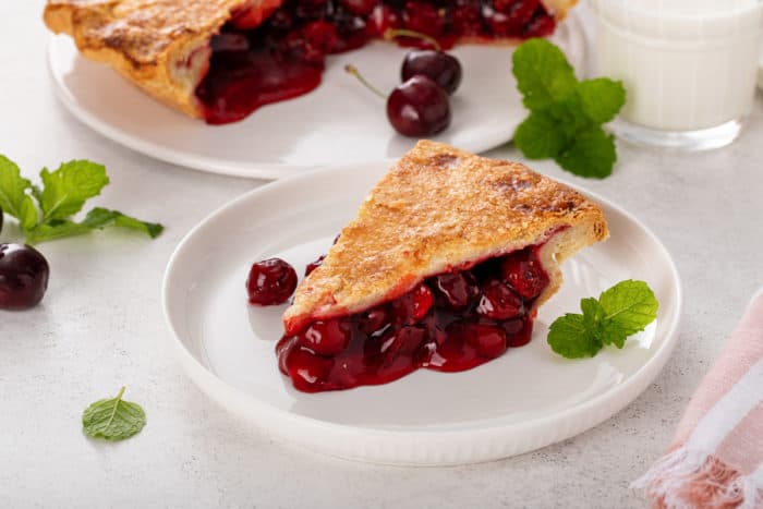 Slice of cherry pie on a white plate. The rest of the pie is just visible in the background.