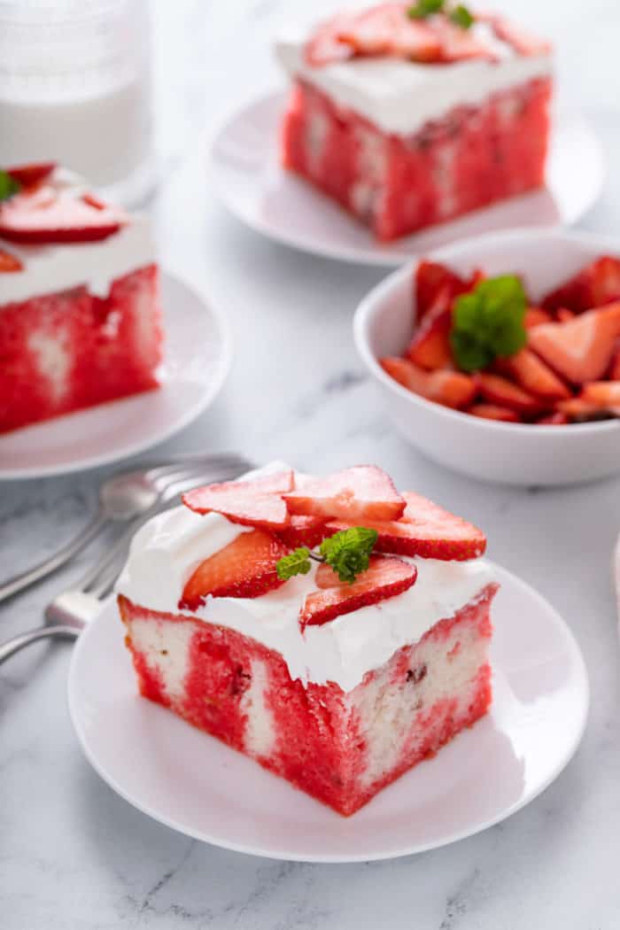 White plate holding a slice of strawberry poke cake. Three more plates of cake and a white bowl filled with sliced strawberries are visible in the background.