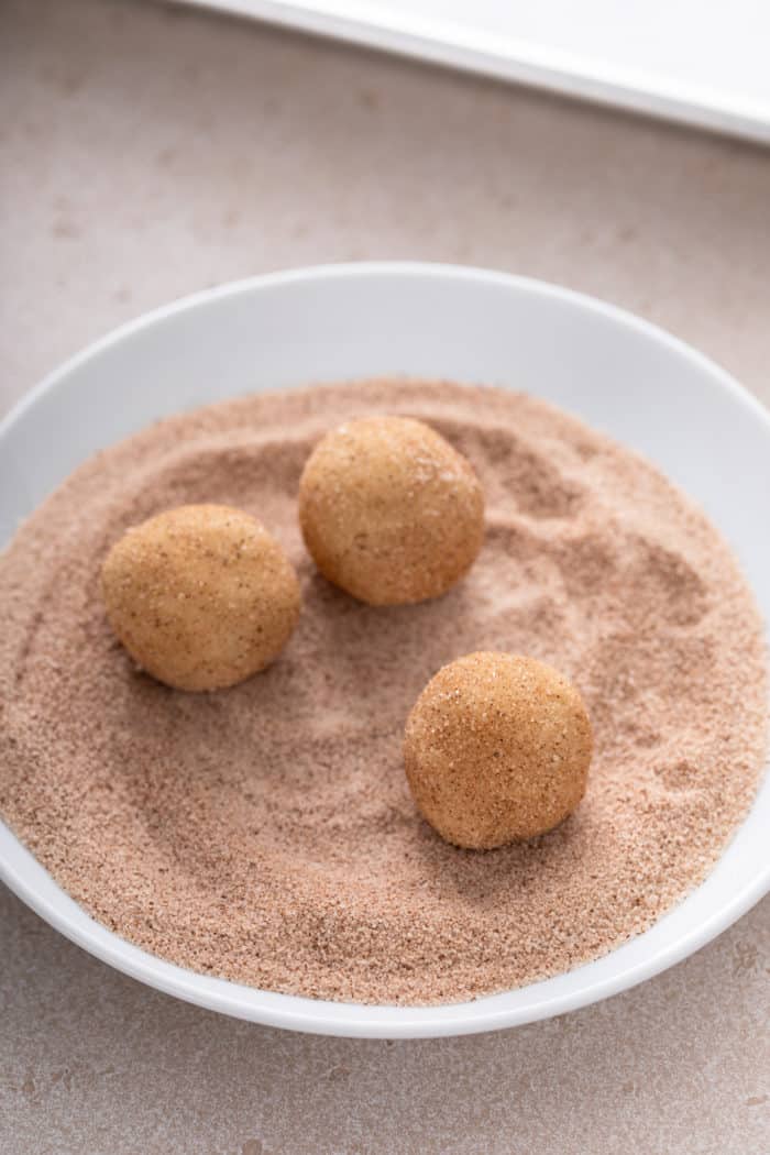 Three cookie dough balls being rolled in a bowl of cinnamon sugar.