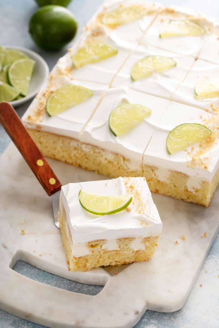 Cake server picking up a slice of key lime poke cake from a white cutting board.