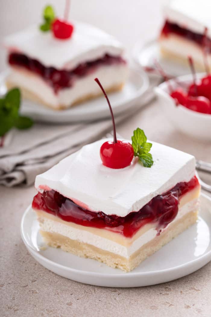 White plate with a slice of layered cherry cheesecake dessert topped with a maraschino cherry and sprig of mint.