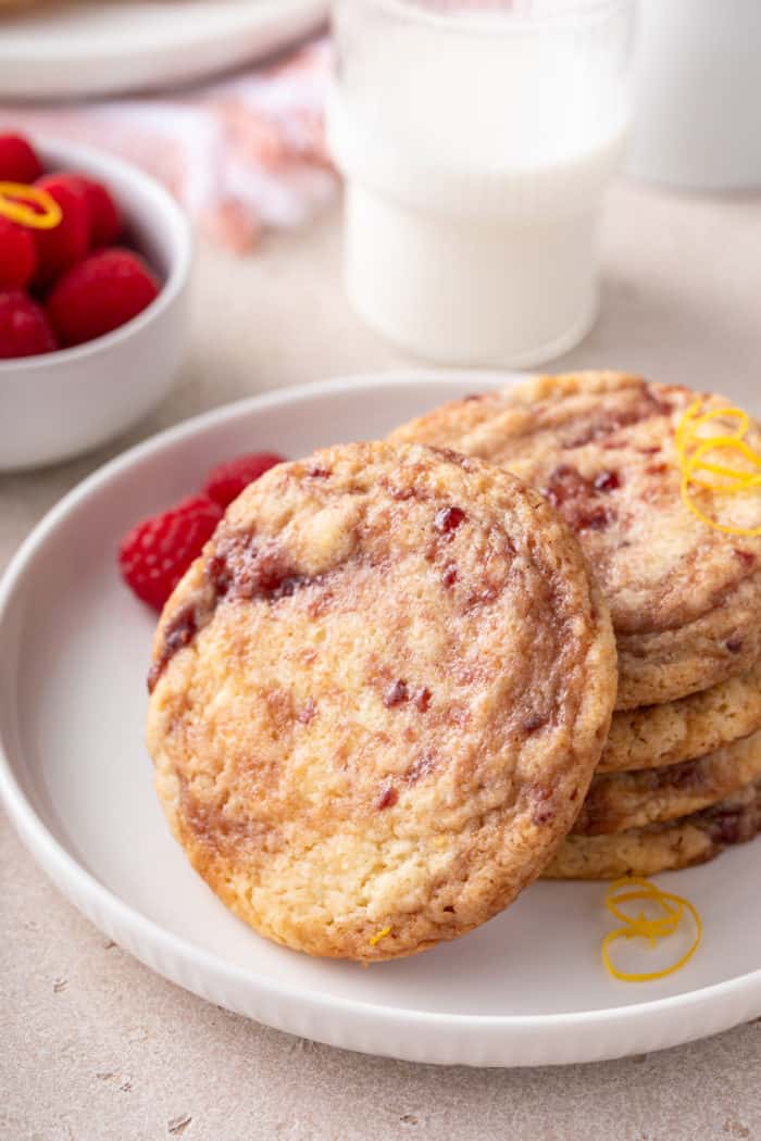 Stack of several raspberry lemon cookies on a white plate. Another raspberry lemon cookie is leaning against the stack.