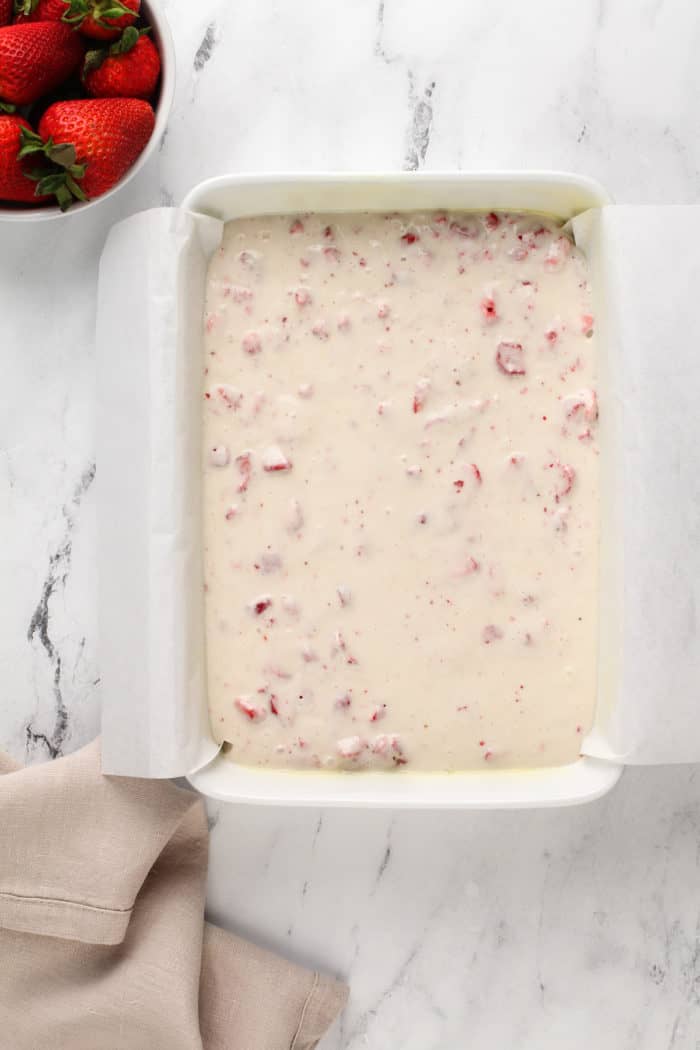 White cake mix with freeze dried strawberries in it spread into a parchment-lined cake pan.