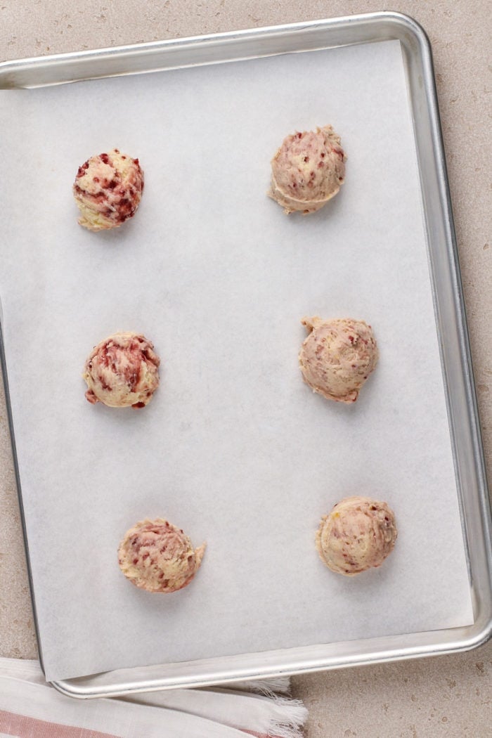 Six large balls of raspberry lemon cookie dough on a lined baking sheet, ready to go in the oven.