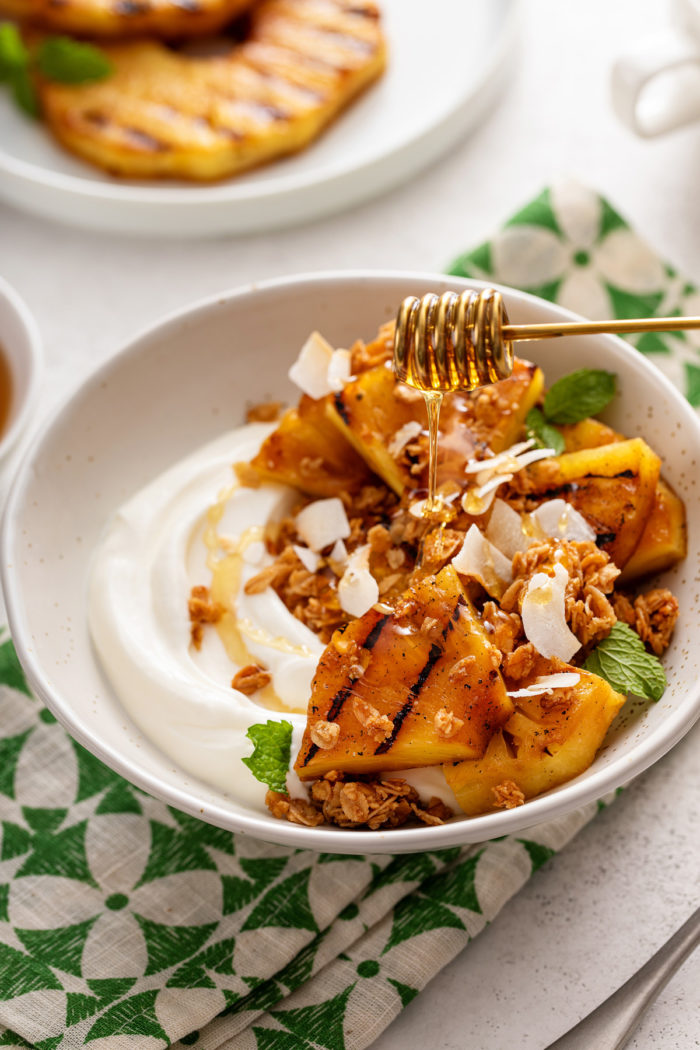 Honey being drizzled over grilled pineapple on yogurt, garnished with coconut.