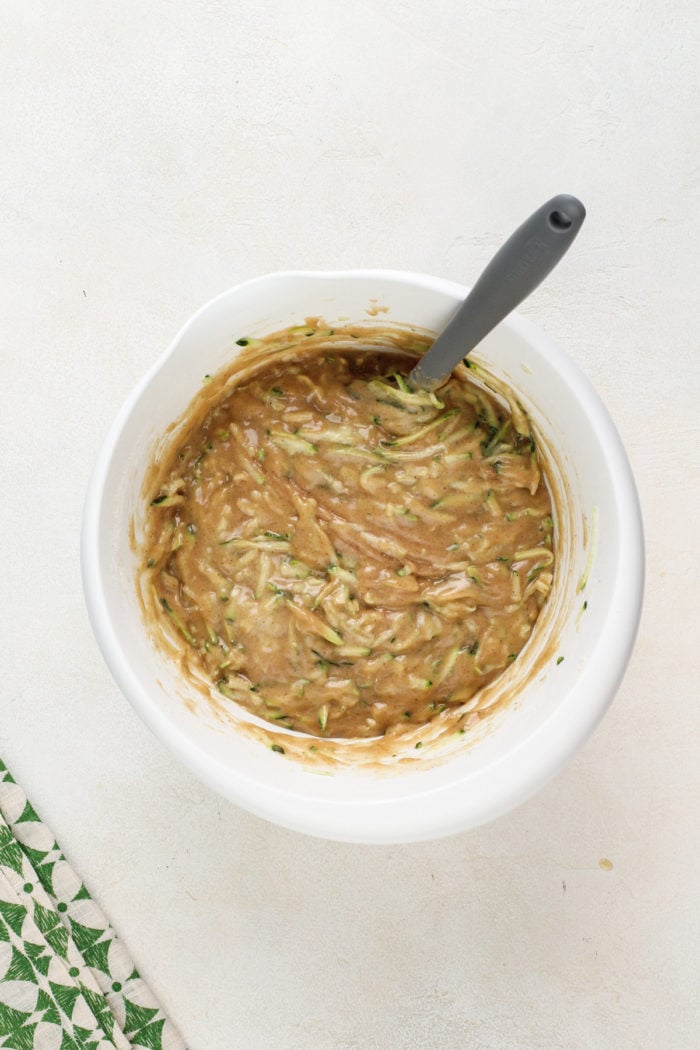 Grated zucchini being folded into muffin batter in a white mixing bowl.