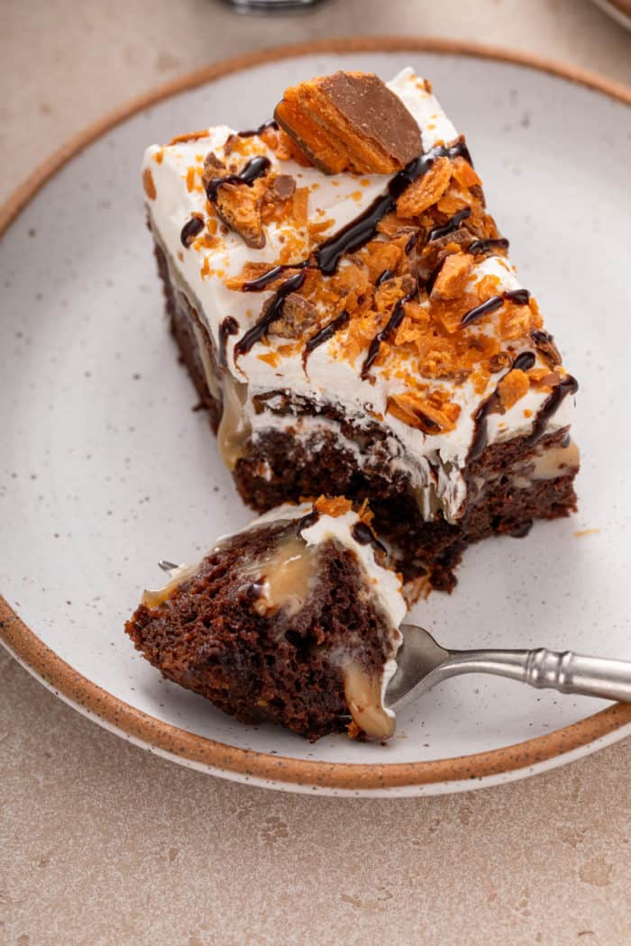 Fork cutting a bite from the corner of a slice of butterfinger poke cake on a plate.