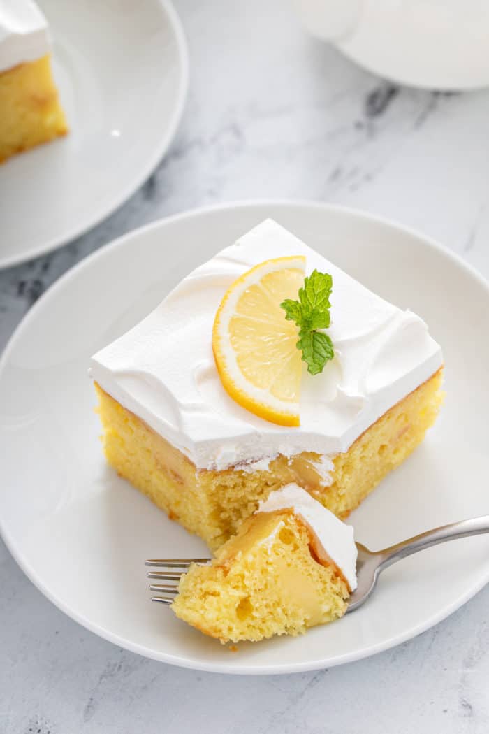 Fork cutting a bite of cake from a slice of lemon poke cake on a white plate.