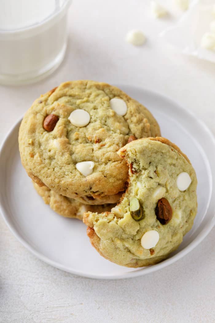 Bite taken from a white chocolate pistachio pudding cookie that is leaning against a few other cookies on a white plate.